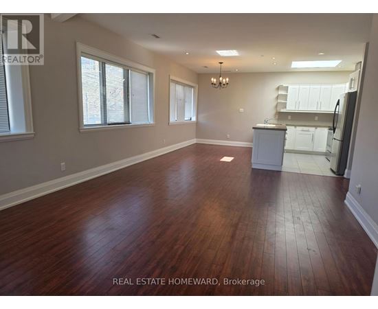 A large empty room with wood flooring and a kitchen in the background.