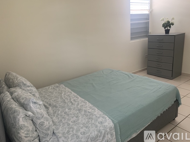 A bed with a grey and white patterned duvet and a grey blanket is in a room with a grey dresser and a window.