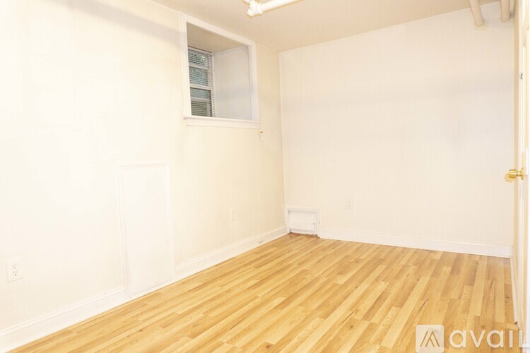 A room with wooden flooring and white walls, with a small window and a door on the right.
