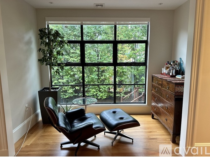 A room with a black chair and a black ottoman in front of a large window with a view of trees outside.
