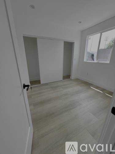 A room with wooden flooring and white walls, with a door on the left and a window on the right.