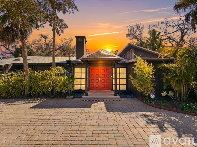 A house with a red door is surrounded by greenery and has a brick walkway leading to it.