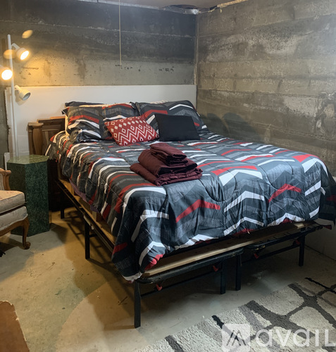 A bed with a grey and red comforter is in a room with concrete walls.