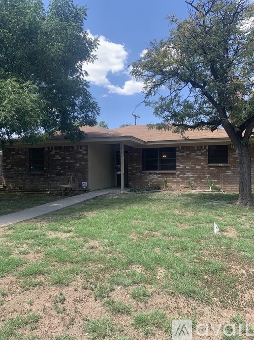 A house with a brown brick exterior and a white door is surrounded by a grassy area and trees.