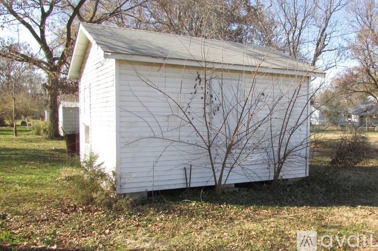 A small white shed with a grey roof is surrounded by trees and grass.