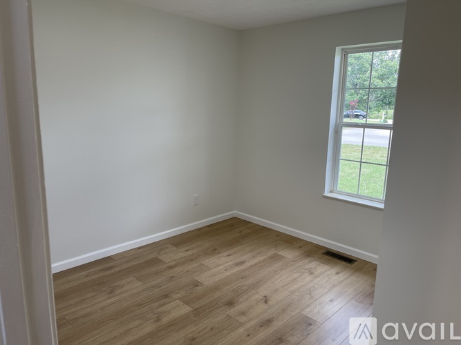 A room with wooden flooring and a window showing a view of a car and trees outside.