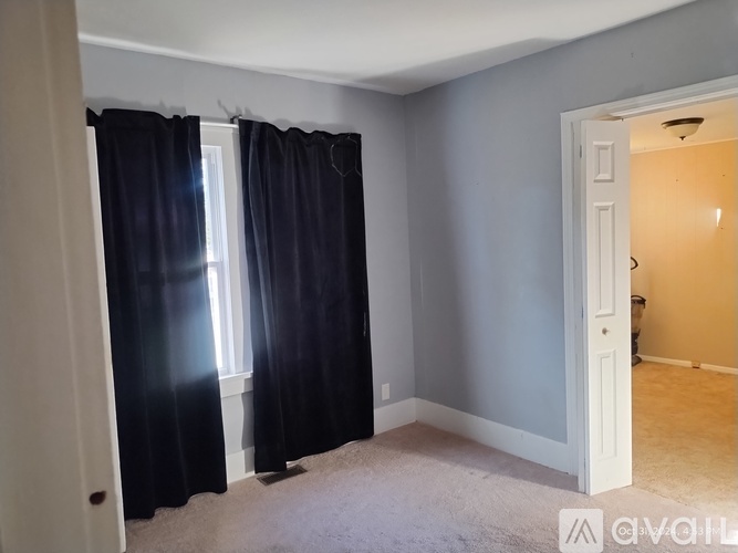 A room with a carpeted floor, a window with black curtains, and a door leading to another room.