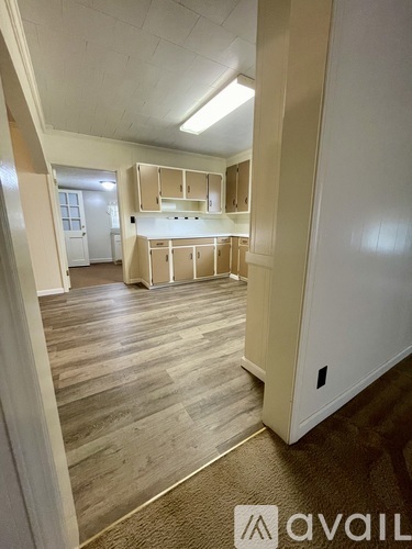 A room with wooden floors and white walls, with a kitchen area in the background.