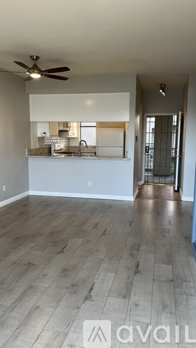A room with a fan and a kitchen area in the background.