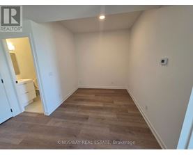 A room with wooden flooring and white walls, with a kitchen visible in the background.