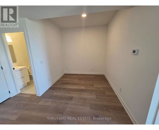 A room with wooden flooring and white walls, with a kitchen visible in the background.