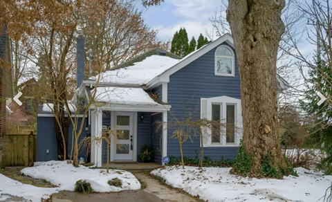 A blue house with a white door and windows, surrounded by trees and snow.
