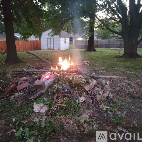 A campfire burns in a backyard surrounded by trees and a fence.