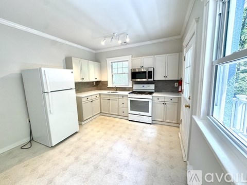 A white refrigerator stands in a kitchen with white cabinets and a window.
