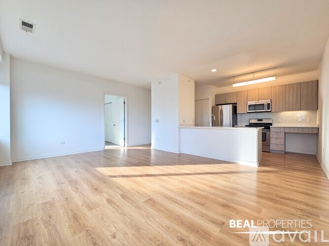 A spacious, empty room with wooden flooring and a kitchen area in the background.