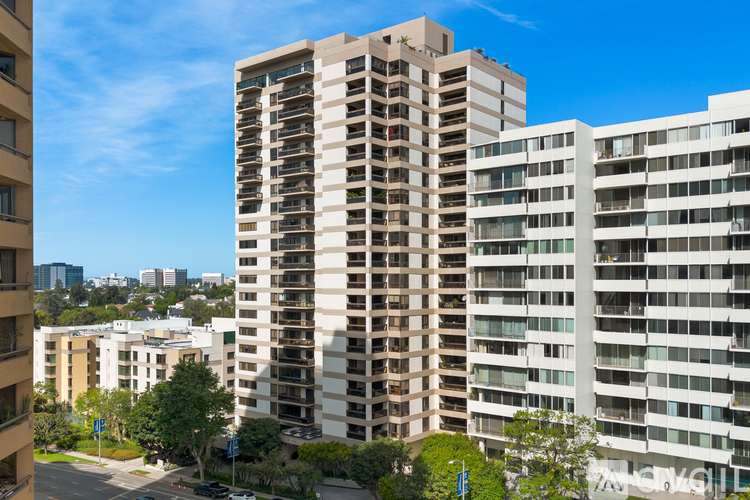 A tall building with balconies is in the foreground with other buildings in the background.