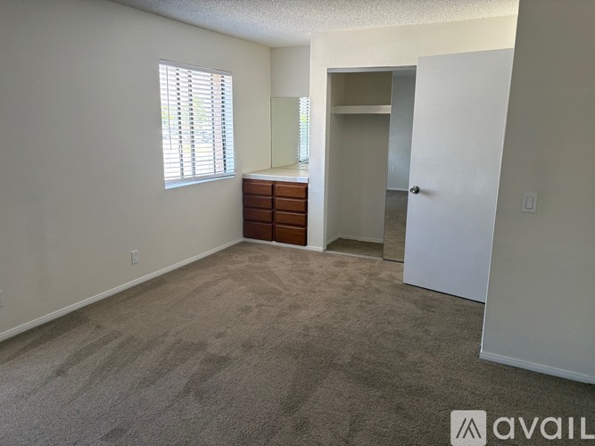 A room with a carpeted floor, a window with blinds, a wooden dresser, and a white door.