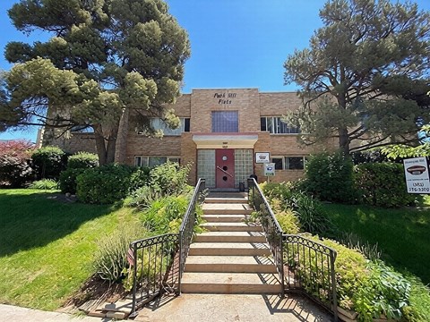 A building with a brick facade and a red door is surrounded by greenery.