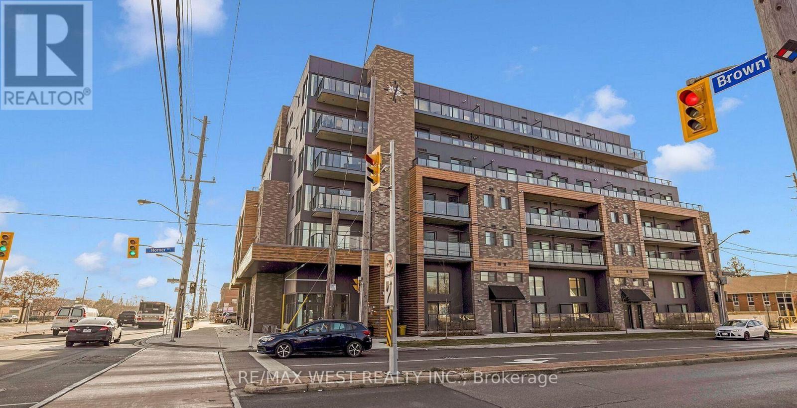 A multi-story building with a brown brick facade is situated on a street corner with traffic lights and cars in the foreground.