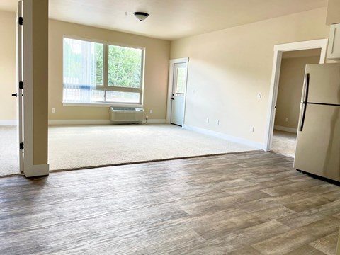 A spacious living room with a large window and a refrigerator in the corner.