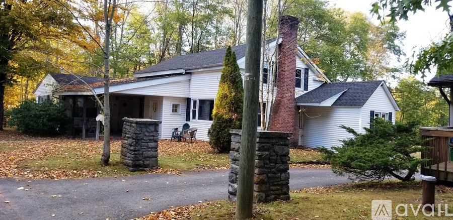 A house with a white exterior and a chimney is surrounded by trees and a stone pillar.