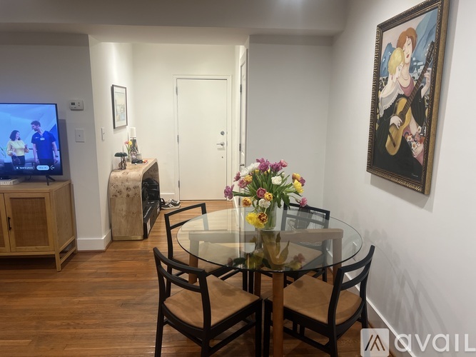 A dining room with a glass table and chairs, a TV, a cabinet, a vase of flowers, and a painting on the wall.