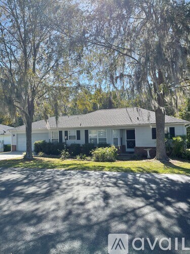 A white house with a grey roof is surrounded by trees and has a driveway in front.
