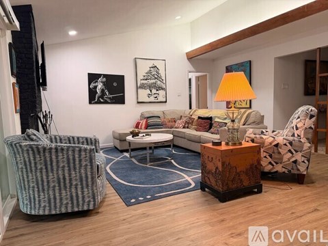 A living room with a grey patterned armchair, a white sofa, a wooden side table with a lamp, and a blue rug.