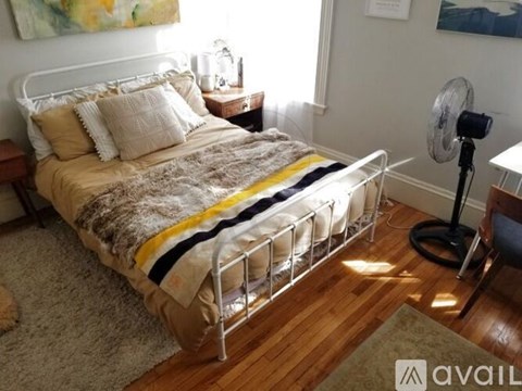 A bed with a white headboard and a yellow and black striped blanket is in the foreground of a room with a fan and a chair.