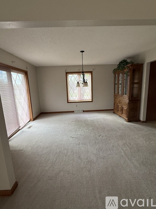 A room with a carpeted floor, a window with blinds, a chandelier, and a wooden cabinet.
