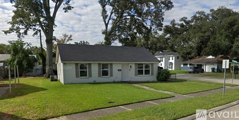 A small white house with a grey roof is surrounded by green grass and trees.