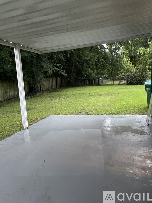 A wet concrete slab under a covered area with trees and a green trash can in the background.