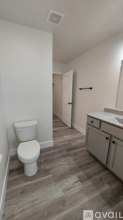 A white toilet sits in a bathroom with wood flooring and a white wall.