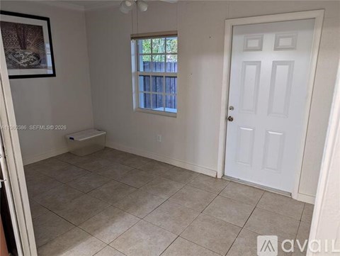 A room with a white door, a window with a view of a fence and trees, and a framed picture on the wall.