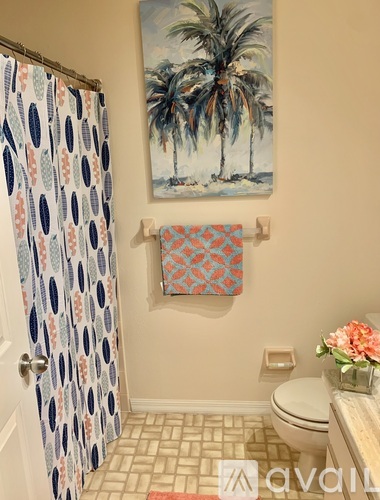 A bathroom with a toilet, a shower curtain, a towel, and a painting of palm trees on the wall.