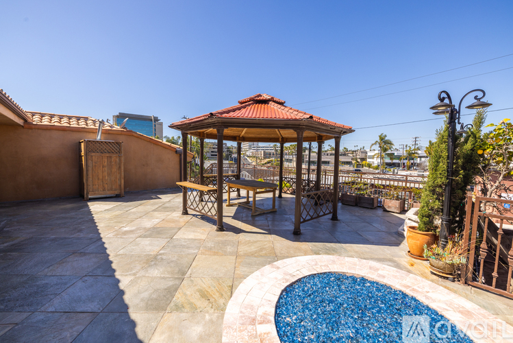 A gazebo with a table and chairs is surrounded by a pool and plants.