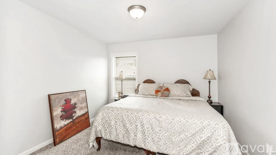 A bedroom with a bed, pillows, a lamp, and a picture of a red flower.
