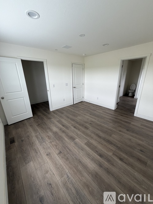 A room with wooden flooring and white walls, with a door on the left and a bathroom visible through the doorway on the right.