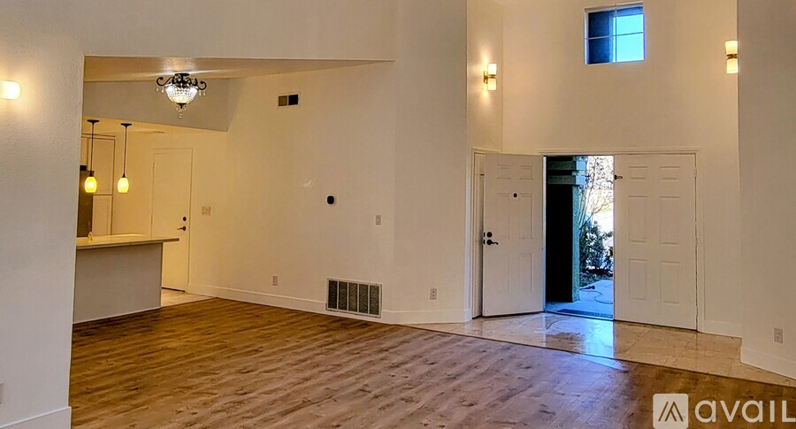A spacious room with wooden floors and white walls, featuring a chandelier, a window, and a door leading outside.
