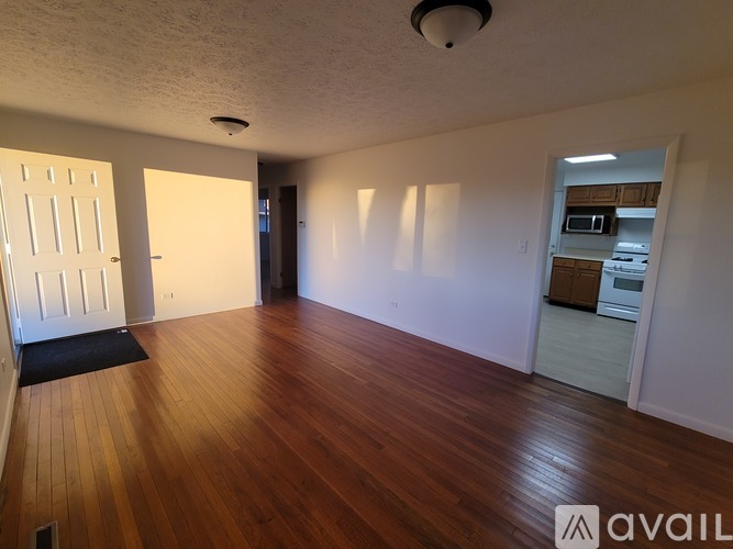A room with wooden floors and white walls, with a door on the left and a kitchen area on the right.