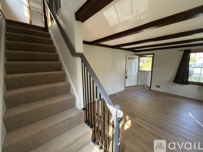 A staircase with a metal railing and beige carpeted steps leads to a spacious room with wooden floors and a large window.