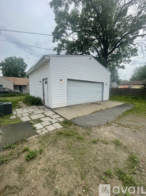 A small white building with a garage door is surrounded by a grassy area and a tree.