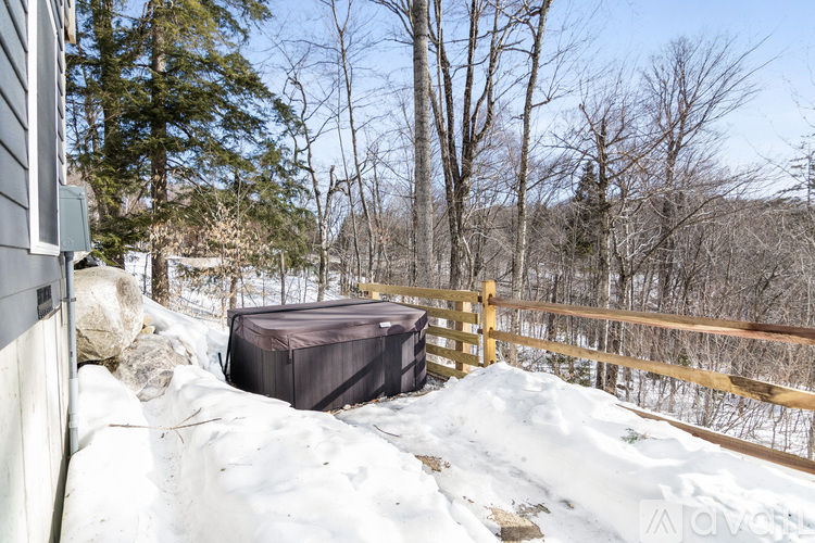 A hot tub sits on a snowy deck with a wooden fence and bare trees in the background.