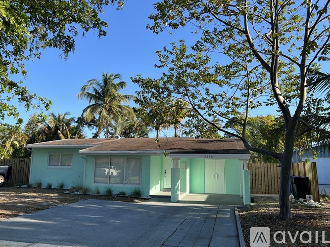 A house with a green exterior is surrounded by trees and has a driveway in front.