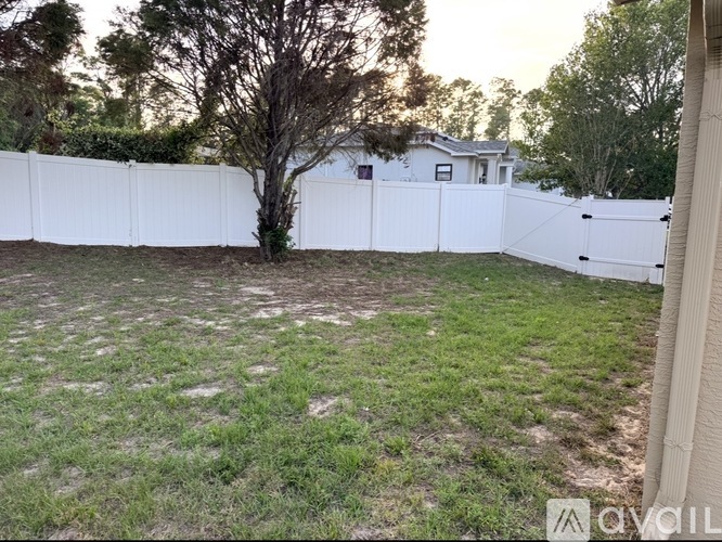 A white fence encloses a grassy yard with a tree and a house in the background.