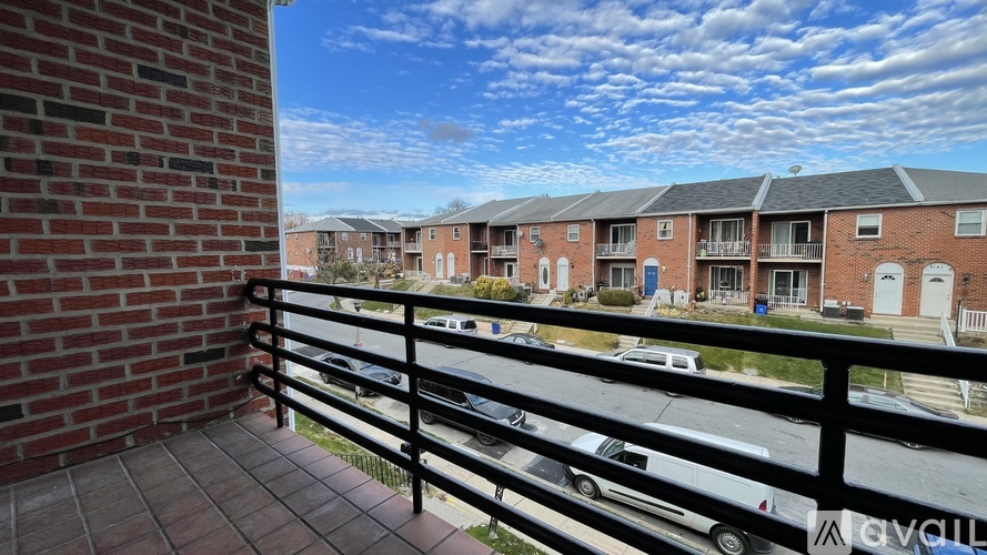 A balcony with a black railing overlooks a street with parked cars and apartment buildings.