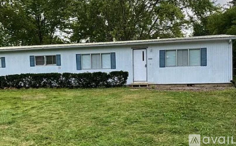 A white mobile home with a white door and windows, surrounded by a green lawn and trees.