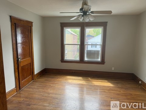 A room with a wooden floor, a ceiling fan, and a window with a view of a building outside.