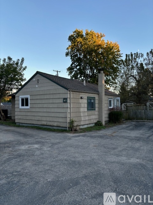 A small house with a grey roof and a chimney is surrounded by a gravel driveway.