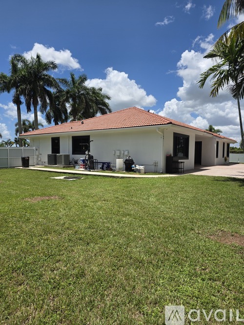 A white house with a red roof is surrounded by a grassy area and palm trees.
